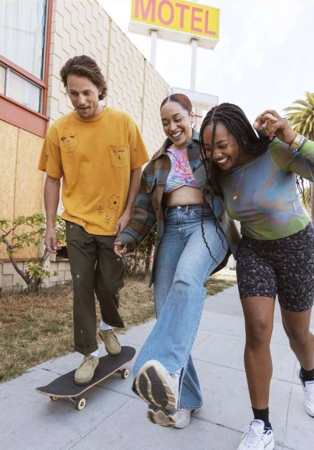 Three young adults walking on a sidewalk with a skateboard near a motel, smiling and posing for the camera in casual clothing.