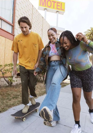 Three young adults walking on a sidewalk with a skateboard near a motel, smiling and posing for the camera in casual clothing.