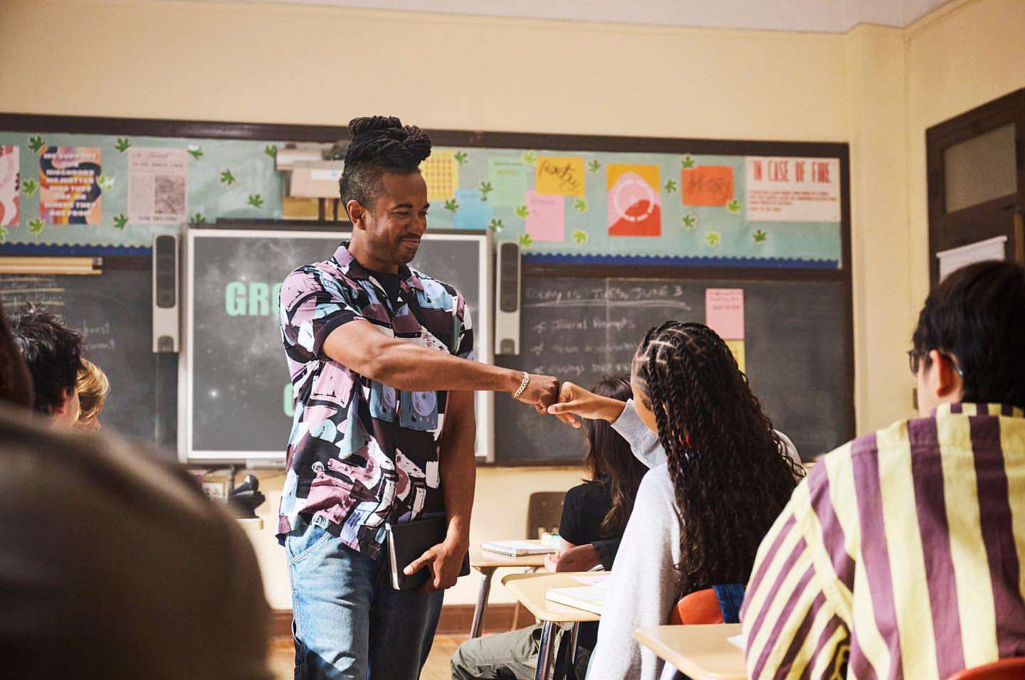 Teacher and student bump fists in a classroom setting. The teacher has dreadlocks and a patterned shirt; the student has long braids.