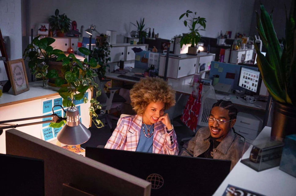 Two smiling professionals, an African-American man and a mixed-race woman, work in a brightly lit office, collaborating on a computer screen.