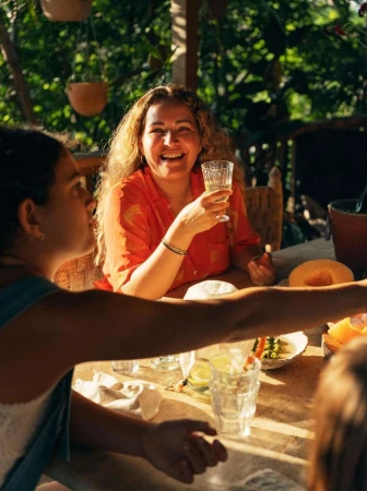 Woman smiles while seated at an outdoor table with friends, holding a drink in a glass, with lush greenery in the background.