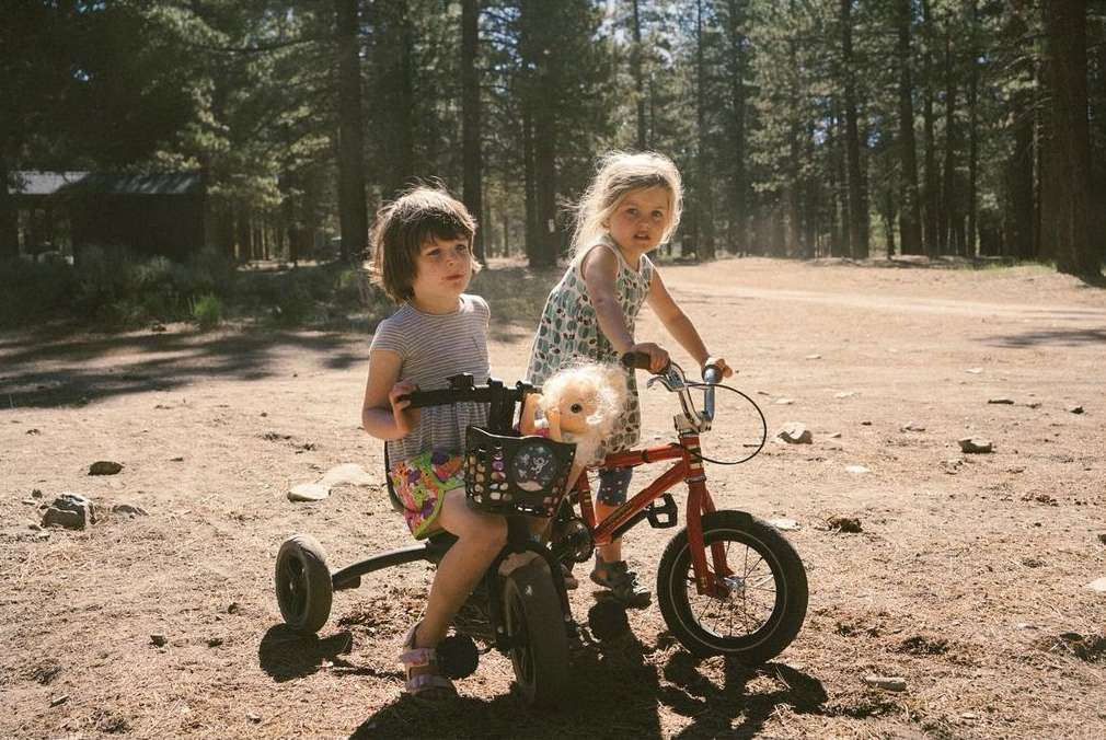 Two young girls on a red bicycle in a forest. One girl sits in the front with a doll, while the other sits behind her.