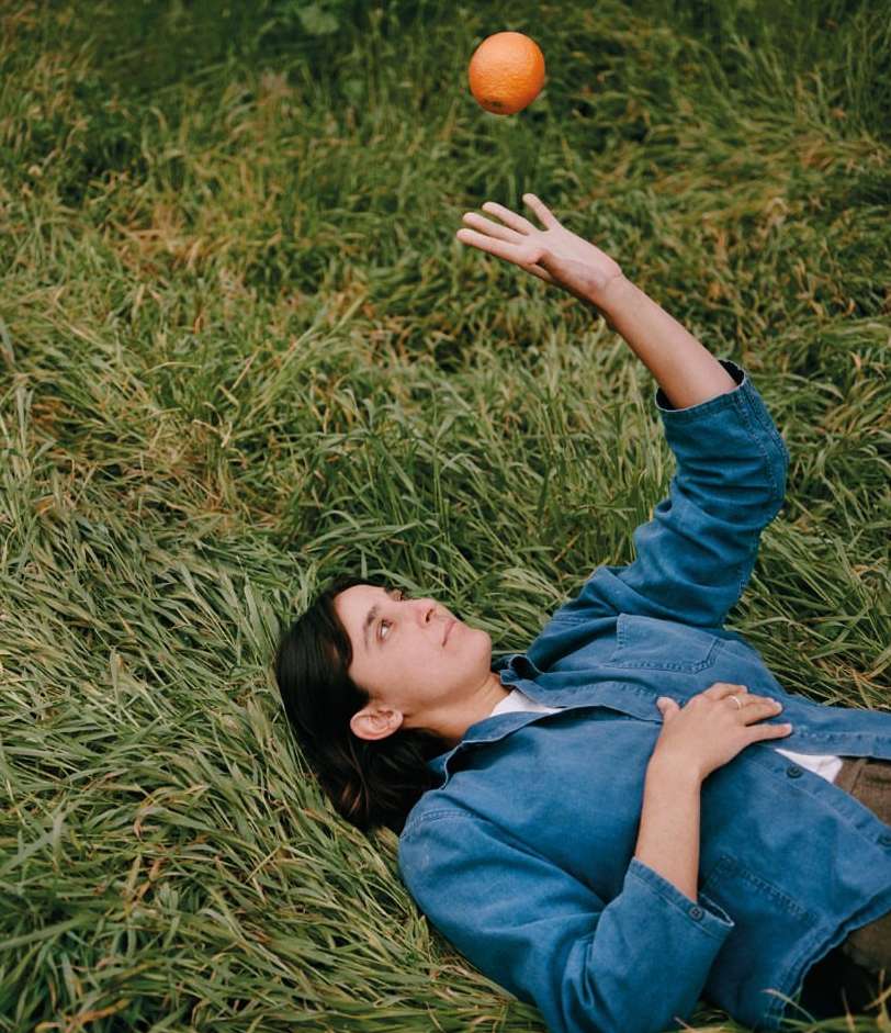 Woman lying on green grass reaches up towards an orange fruit floating above her hand in outdoor setting during daytime.