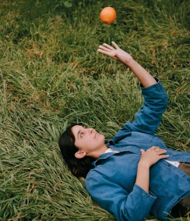 Woman lying on green grass reaches up towards an orange fruit floating above her hand in outdoor setting during daytime.