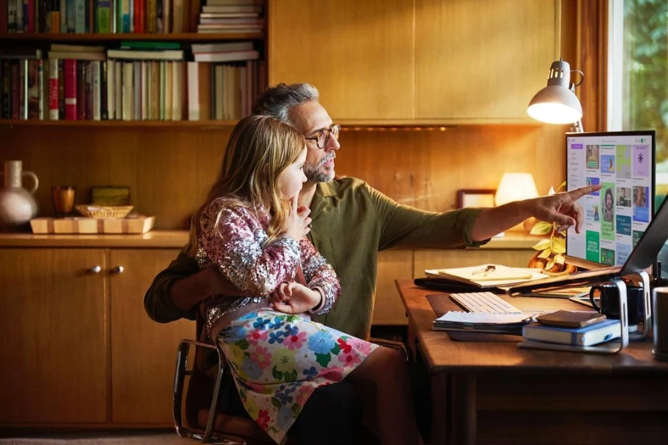 Father and daughter look at a laptop screen with website previews in a home office with warm lighting and bookshelves in the background.