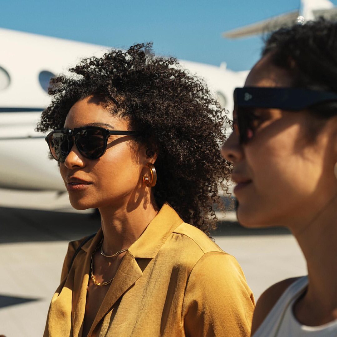 Two adult women stand in front of a private jet with blue sky. One woman wears a tan blazer, gold hoops, sunglasses and a gold chain necklace.