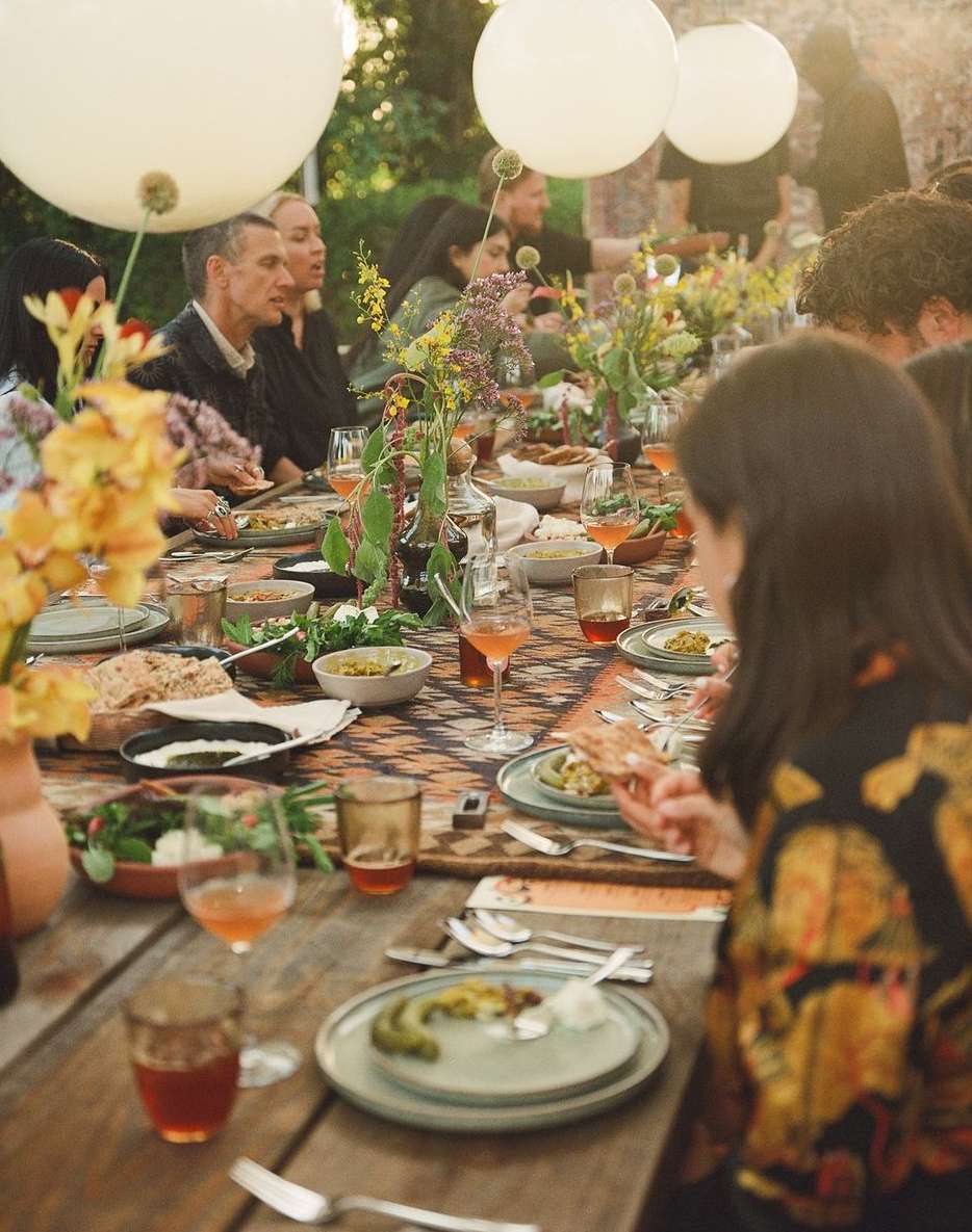 People sit at an outdoor dining table laden with food, drinks, and flowers, illuminated by hanging white balloons in natural light.