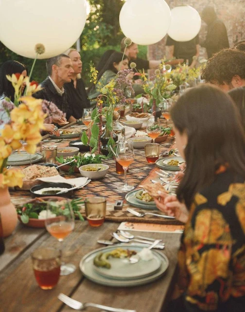 People sit at an outdoor dining table laden with food, drinks, and flowers, illuminated by hanging white balloons in natural light.