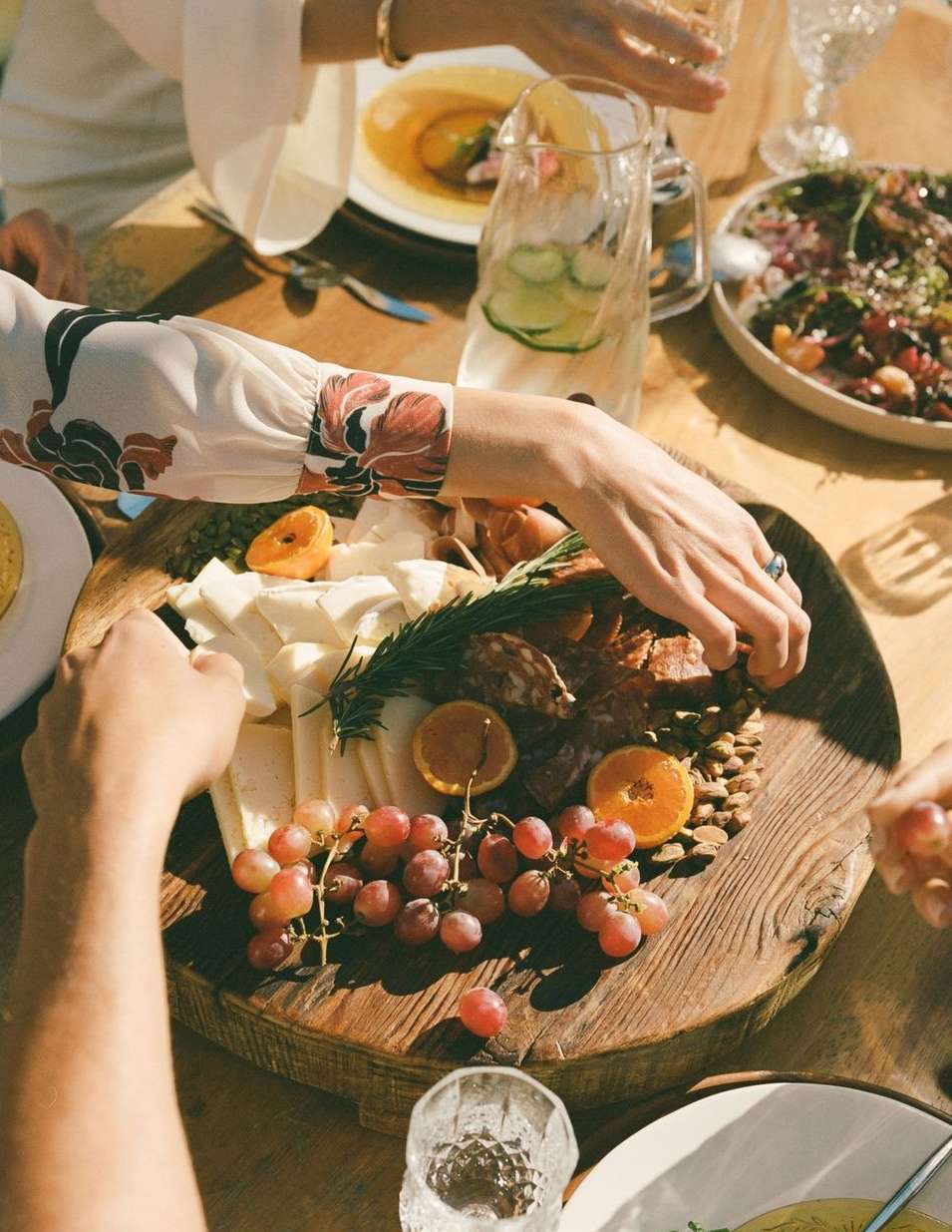 Overhead shot of a wood charcuterie board with cheese, grapes, meats, and citrus, surrounded by people at an outdoor dinner setting.