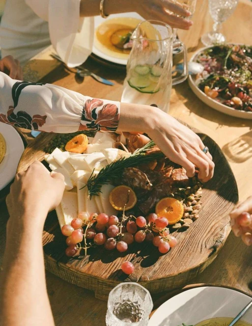 Overhead shot of a wood charcuterie board with cheese, grapes, meats, and citrus, surrounded by people at an outdoor dinner setting.
