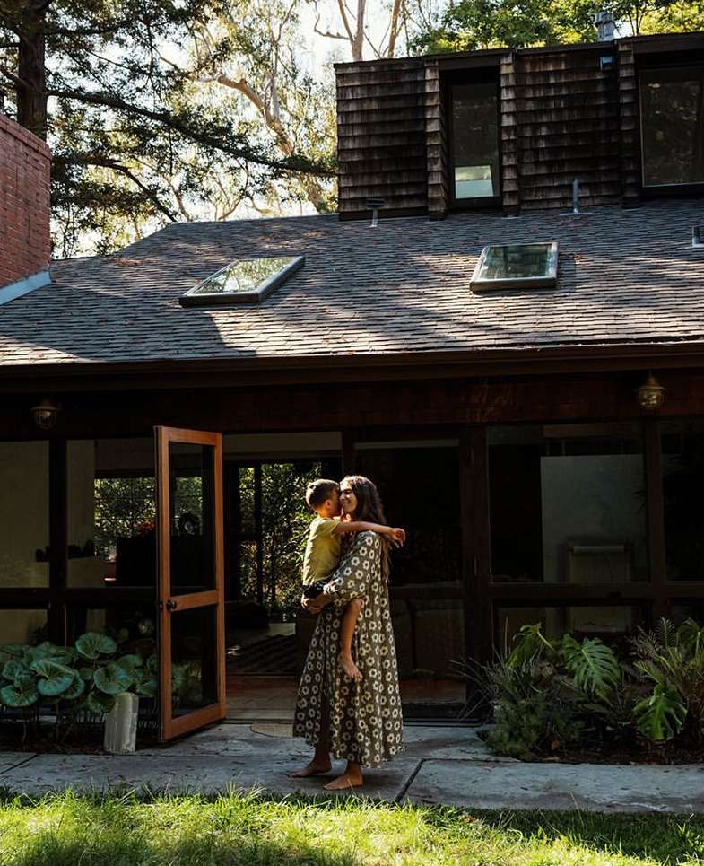 A woman holds a child outside a modern house with a brown roof. The yard is green with plants and trees in a daytime shot.