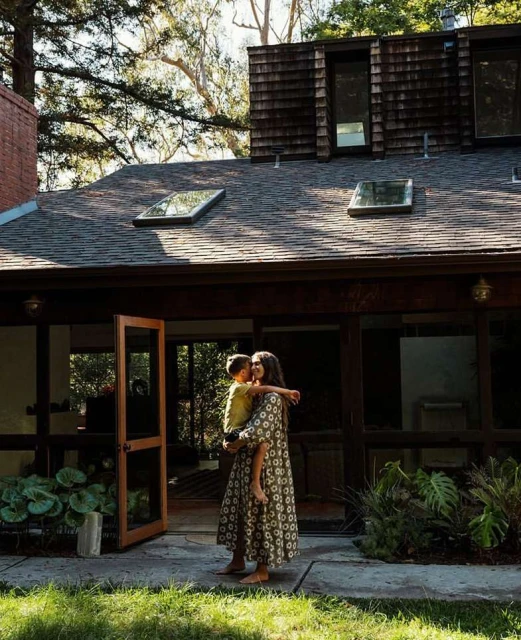 A woman holds a child outside a modern house with a brown roof. The yard is green with plants and trees in a daytime shot.