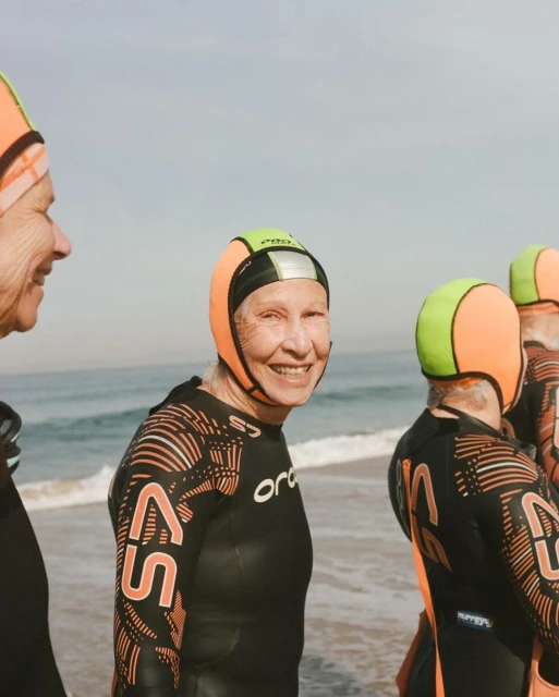 A group of elderly women in wetsuits and swim caps stand on a sandy beach near the ocean, smiling and ready for a swim.