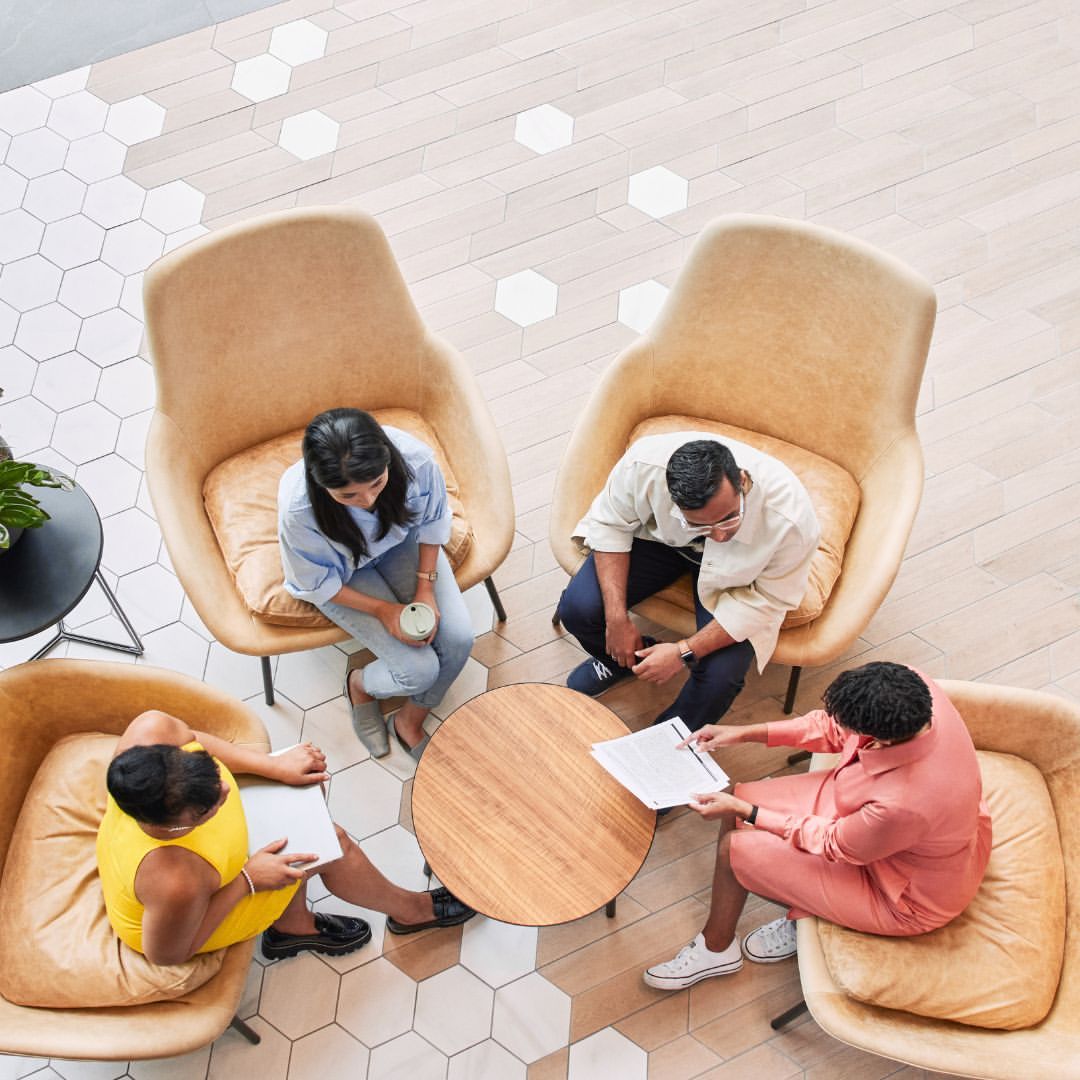 Four adults in a circle of tan chairs collaborate in a well-lit indoor setting, with a modern geometric floor pattern.