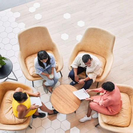 Four adults in a circle of tan chairs collaborate in a well-lit indoor setting, with a modern geometric floor pattern.