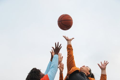 A low angle captures two adults of different ethnicities leaping to catch a basketball against a bright, neutral sky backdrop.