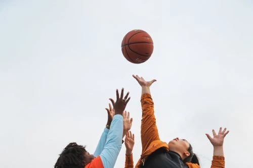 A low angle captures two adults of different ethnicities leaping to catch a basketball against a bright, neutral sky backdrop.