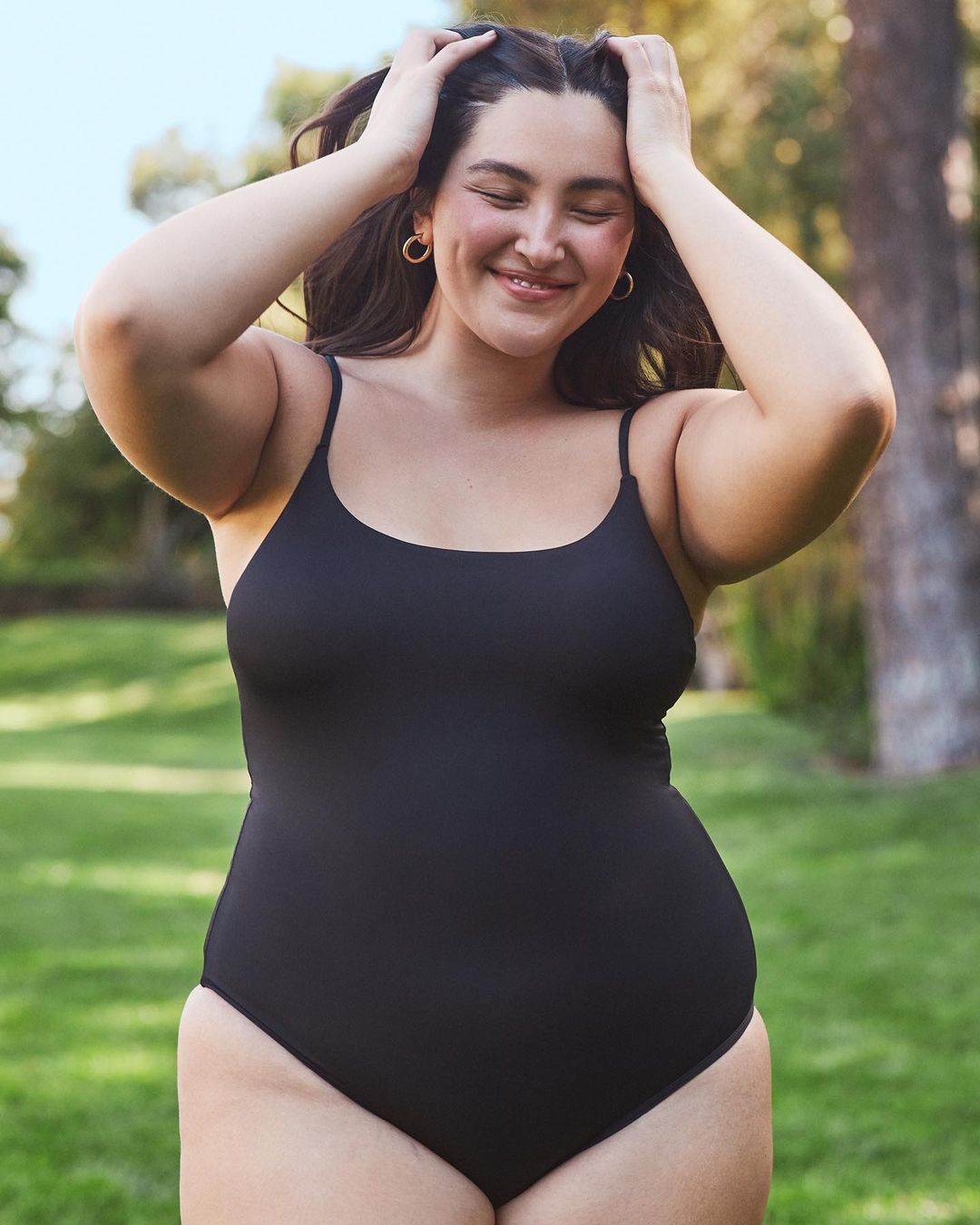Smiling woman in a black one-piece swimsuit poses outdoors with her hands on her head on a sunny day with a grassy background.