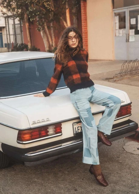 A young woman leans against a white vintage car, wearing a fuzzy checkered sweater and patchwork jeans, set outdoors in natural light.