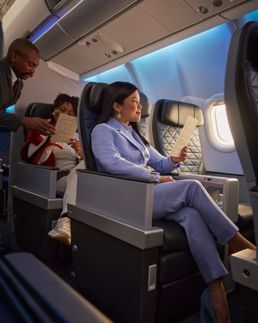 Woman in a blue suit sits in an airplane seat reading a menu with an attendant assisting her. The interior is brightly lit.