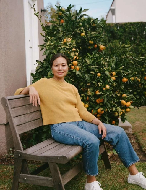 An Asian woman sits on a wooden bench in a backyard next to a citrus tree filled with oranges. She wears a yellow sweater and jeans.