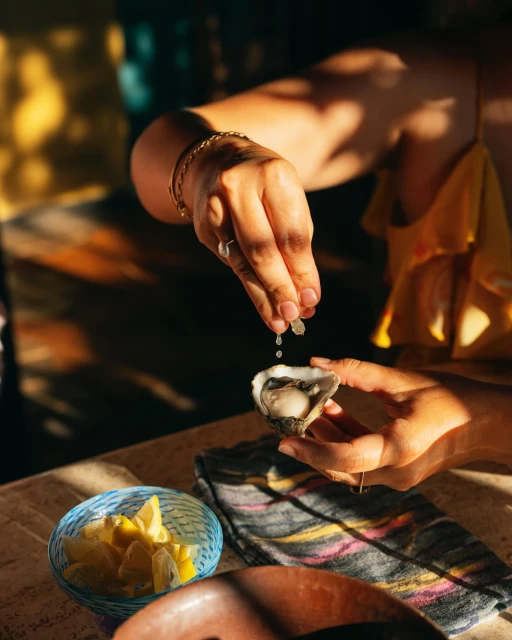 Woman squeezes lemon juice onto an oyster held in her hands, with a bowl of sliced lemons in the background, warm lighting.