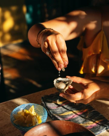 Woman squeezes lemon juice onto an oyster held in her hands, with a bowl of sliced lemons in the background, warm lighting.