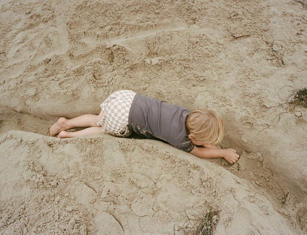 Child lies on sand in checkerboard shorts with a grey shirt on the beach. Blond hair visible and sandy texture is dominant.