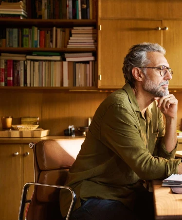 Profile portrait of a mature man with gray hair and glasses sitting at a desk in a home office with a wooden bookshelf.