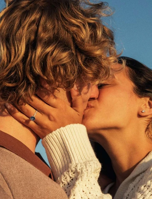 Close-up of a woman's face as she kisses a man, her hand with a ring on his face, with a blue sky in the background.