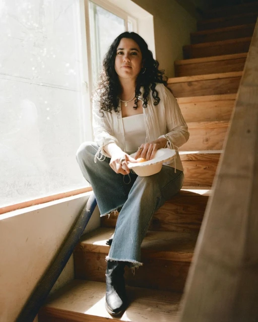 Young woman with curly hair sits on wooden stairs near a sunny window indoors, wearing casual clothing and holding a bowl.