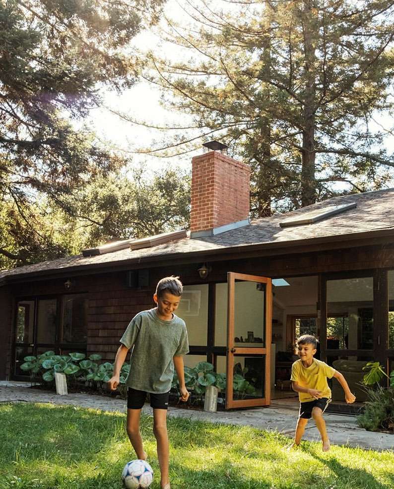 Two boys playing soccer on the lawn in front of a modern home with trees and a brick chimney during the daytime on a sunny day.