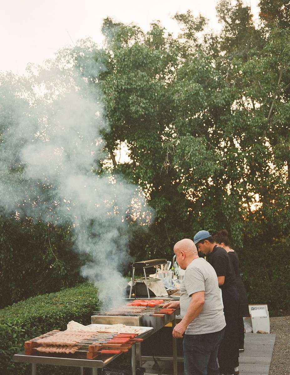 Outdoor barbecue scene with several adults cooking food on grills, smoke rising, with green trees visible in the background, and overcast sky.