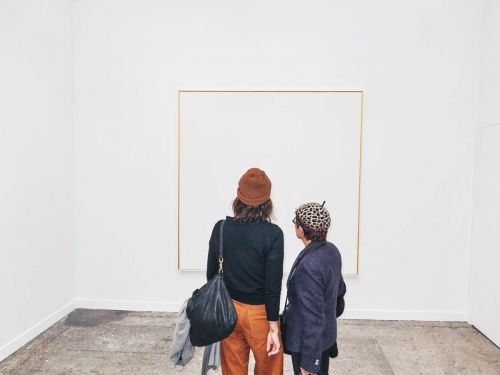 Two people stand in an art gallery looking at a framed white canvas. One wears a brown beanie, the other a patterned hat. Light grey floor.