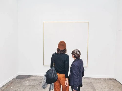 Two people stand in an art gallery looking at a framed white canvas. One wears a brown beanie, the other a patterned hat. Light grey floor.
