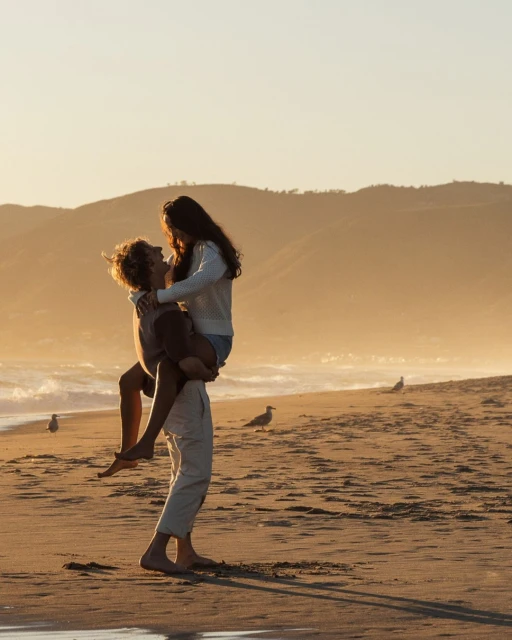 A barefoot couple embraces on a sandy beach with gentle waves and distant mountains during a warm sunset, creating a romantic scene.