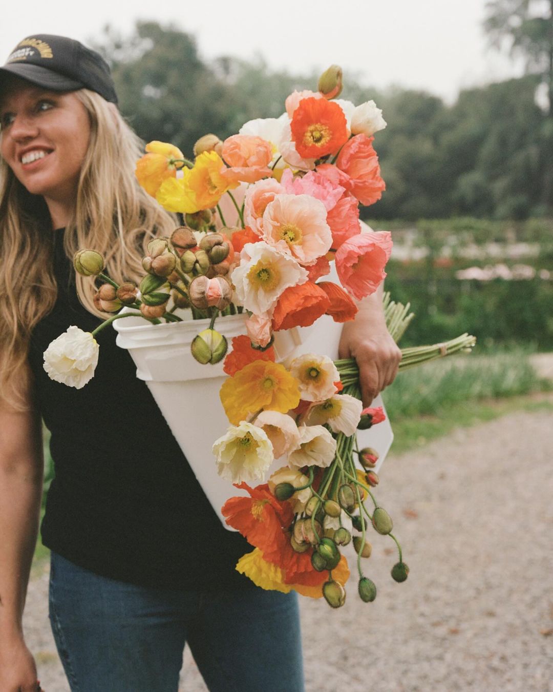 Smiling woman holding a large bucket filled with orange, yellow, and white poppy flowers in a field on a cloudy day.