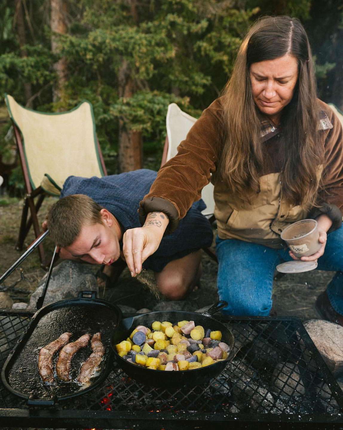 Two people cooking outdoors over a grill, preparing breakfast with potatoes and bacon. Outdoor cooking, nature, and camp breakfast.