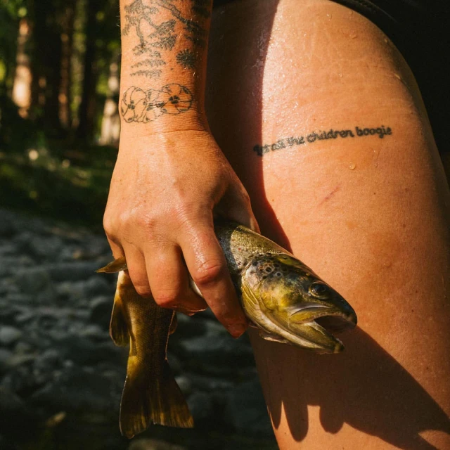Close-up of a person's hand holding a brown trout, with arm tattoos visible, set against a blurred background of trees and foliage.