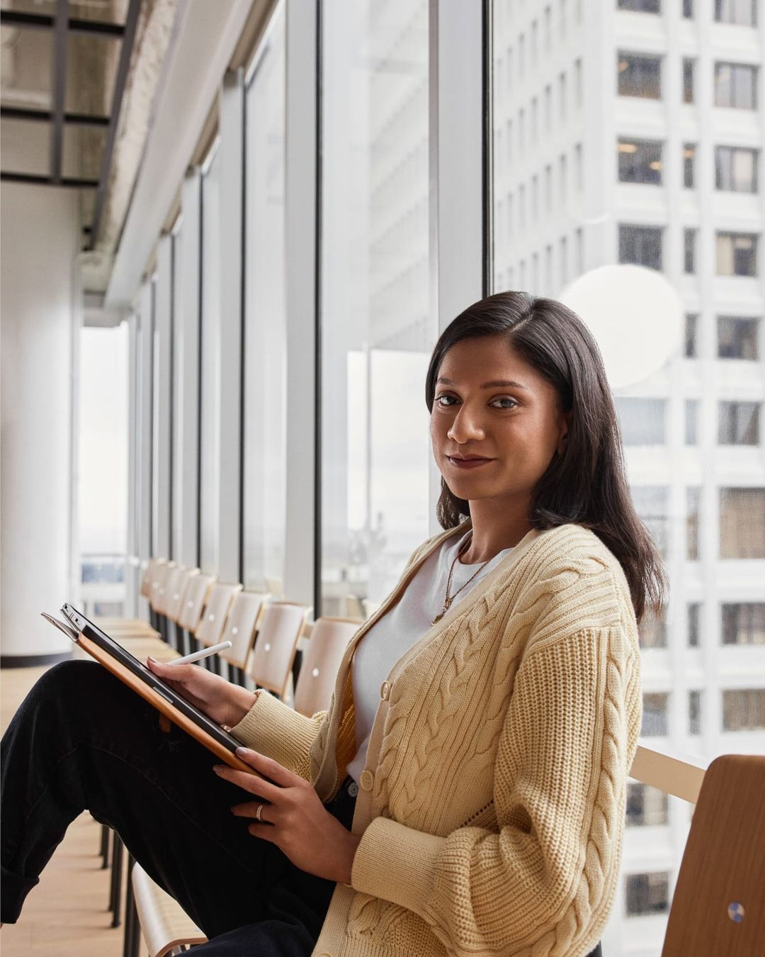 Young woman sits in an office interior with large windows, holding a tablet with a stylus, wearing a cardigan sweater.