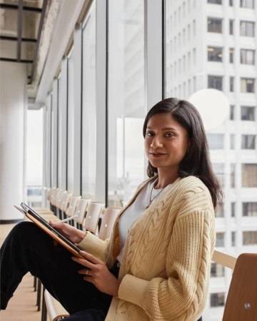 Young woman sits in an office interior with large windows, holding a tablet with a stylus, wearing a cardigan sweater.