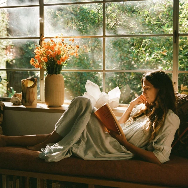 Woman relaxing by window reading a book with orange flowers, quartz crystal, and foliage, creating a serene and intellectual atmosphere.