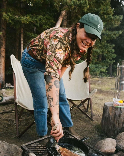 Woman in floral shirt cooks outdoors over fire with a cast iron pan, wearing a baseball cap, near trees and camping chairs.