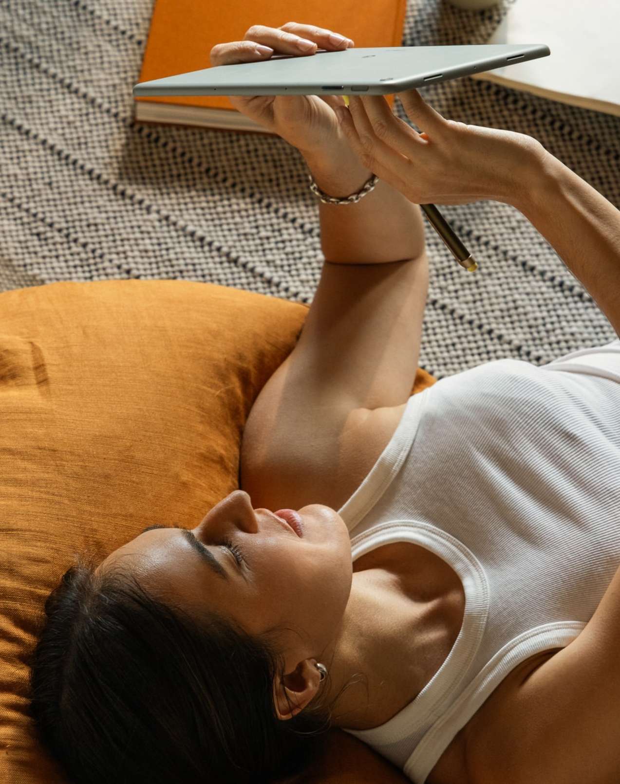 Woman lies on pillow, holds tablet computer, and uses stylus pen. Books, neutral rug, and indoor natural lighting fill frame.