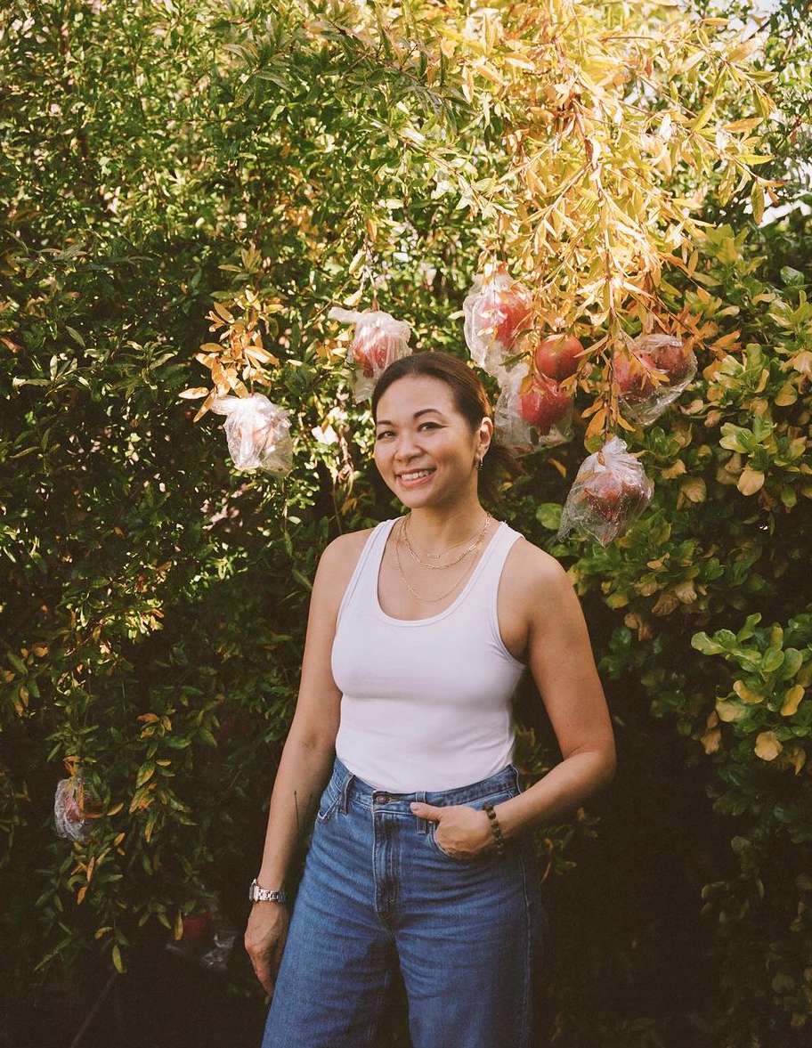 Smiling woman stands in front of a tree with pomegranates; she wears a white tank top, blue jeans, and a wristwatch. Green foliage surrounds her.