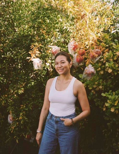 Smiling woman stands in front of a tree with pomegranates; she wears a white tank top, blue jeans, and a wristwatch. Green foliage surrounds her.
