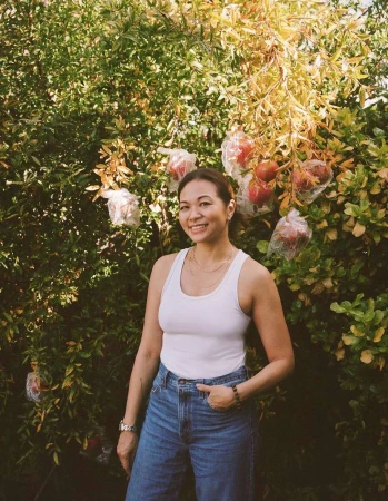 Smiling woman stands in front of a tree with pomegranates; she wears a white tank top, blue jeans, and a wristwatch. Green foliage surrounds her.