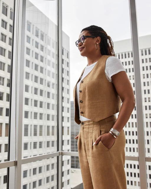 Professional woman with dreadlocks in stylish vest and pants, standing near a window with a city view, wearing glasses and bracelets.