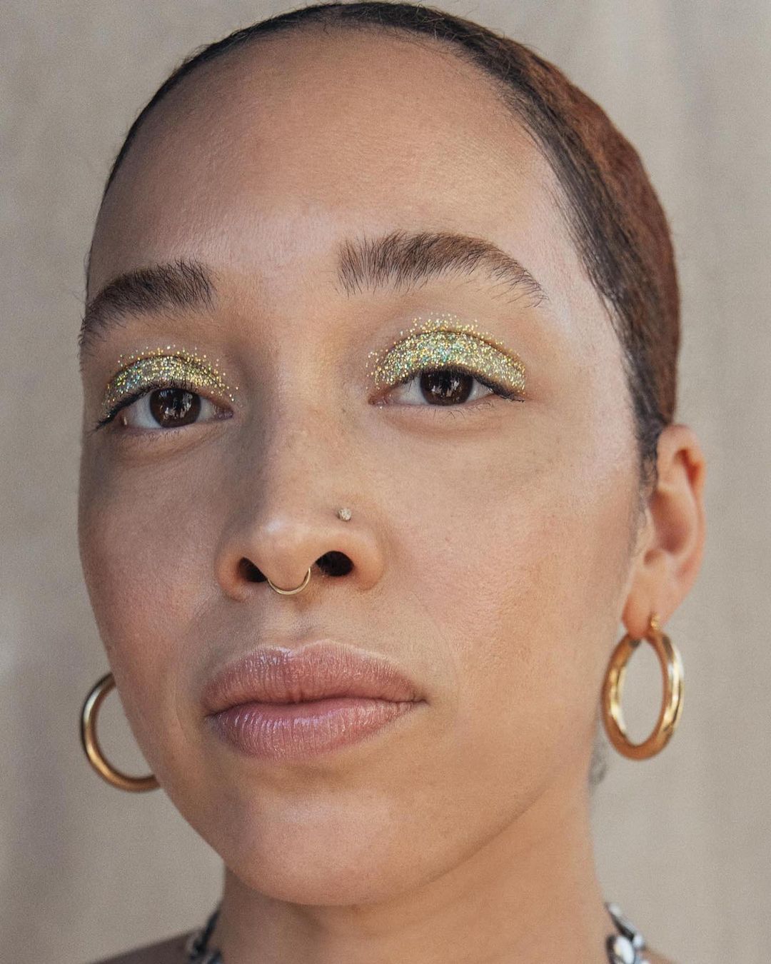 Close-up portrait of a woman with light skin and glittery eyeshadow wearing gold hoop earrings against a neutral background.