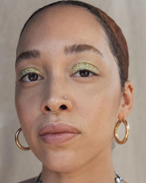 Close-up portrait of a woman with light skin and glittery eyeshadow wearing gold hoop earrings against a neutral background.